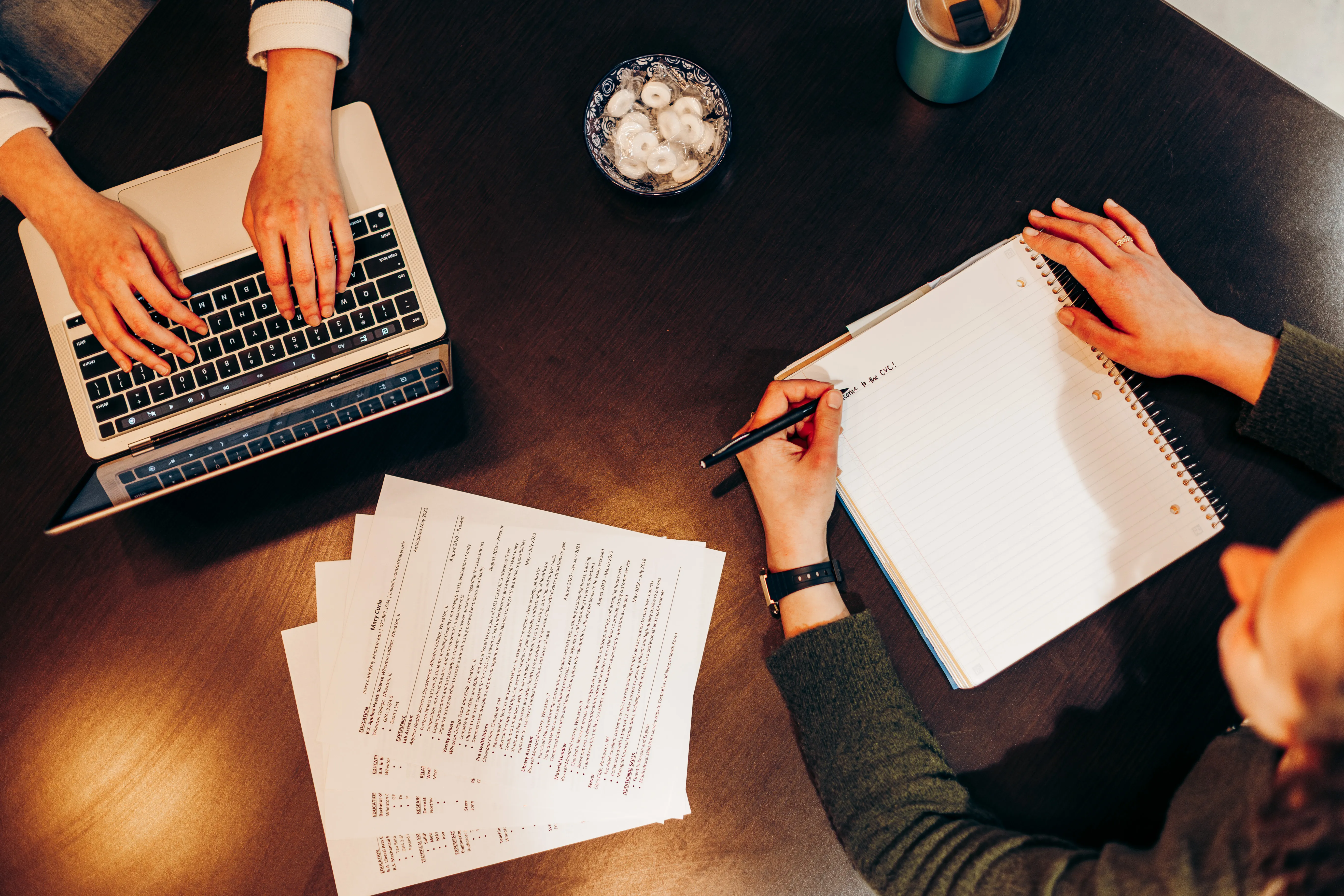 A table with copies of a resume, a notebook, and a computer