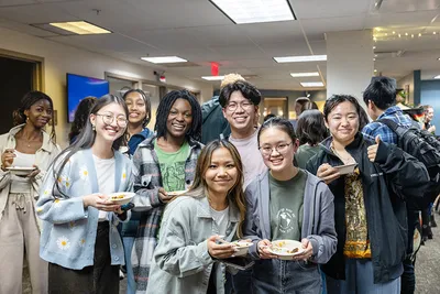 A group of students smiling at the camera and holding plates of food during a social gathering
