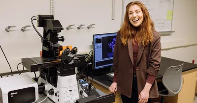 A smiling woman with ginger hair stands to the right of a microscope in a lab.