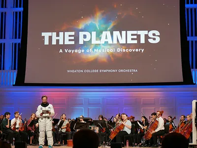 A man in a spacesuit stands in front and an orchestra performs behind him