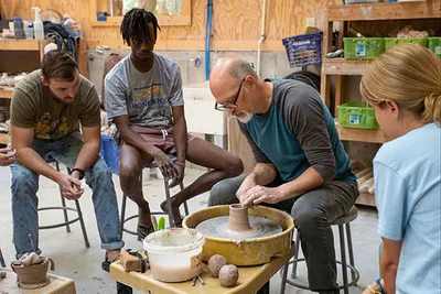 A man demonstrates pottery techniques on a wheel to a group of people in a workshop
